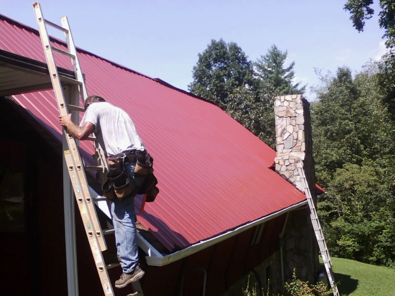 Roofer on a ladder beside a red metal roof and stone chimney — Soffit & Fascia Repair by Roofing Brothers in Bishopville